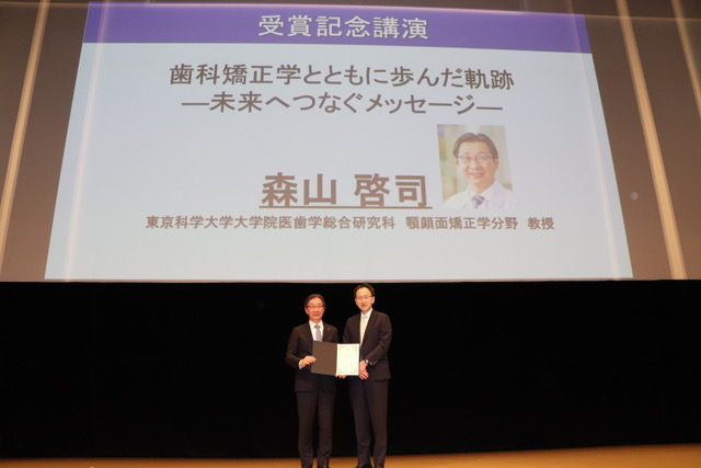 受賞記念講演のスライドの前で賞状を手にする森山教授ら